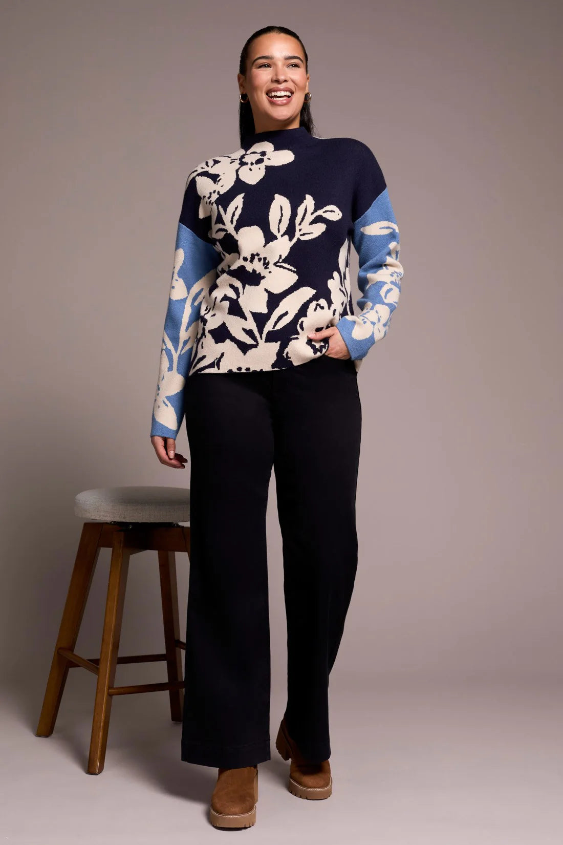 Woman wearing a navy sweater with white floral patterns, standing next to a wooden stool against a plain background.