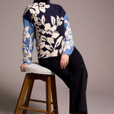 Woman wearing a floral-patterned sweater sitting on a wooden stool against a plain background
