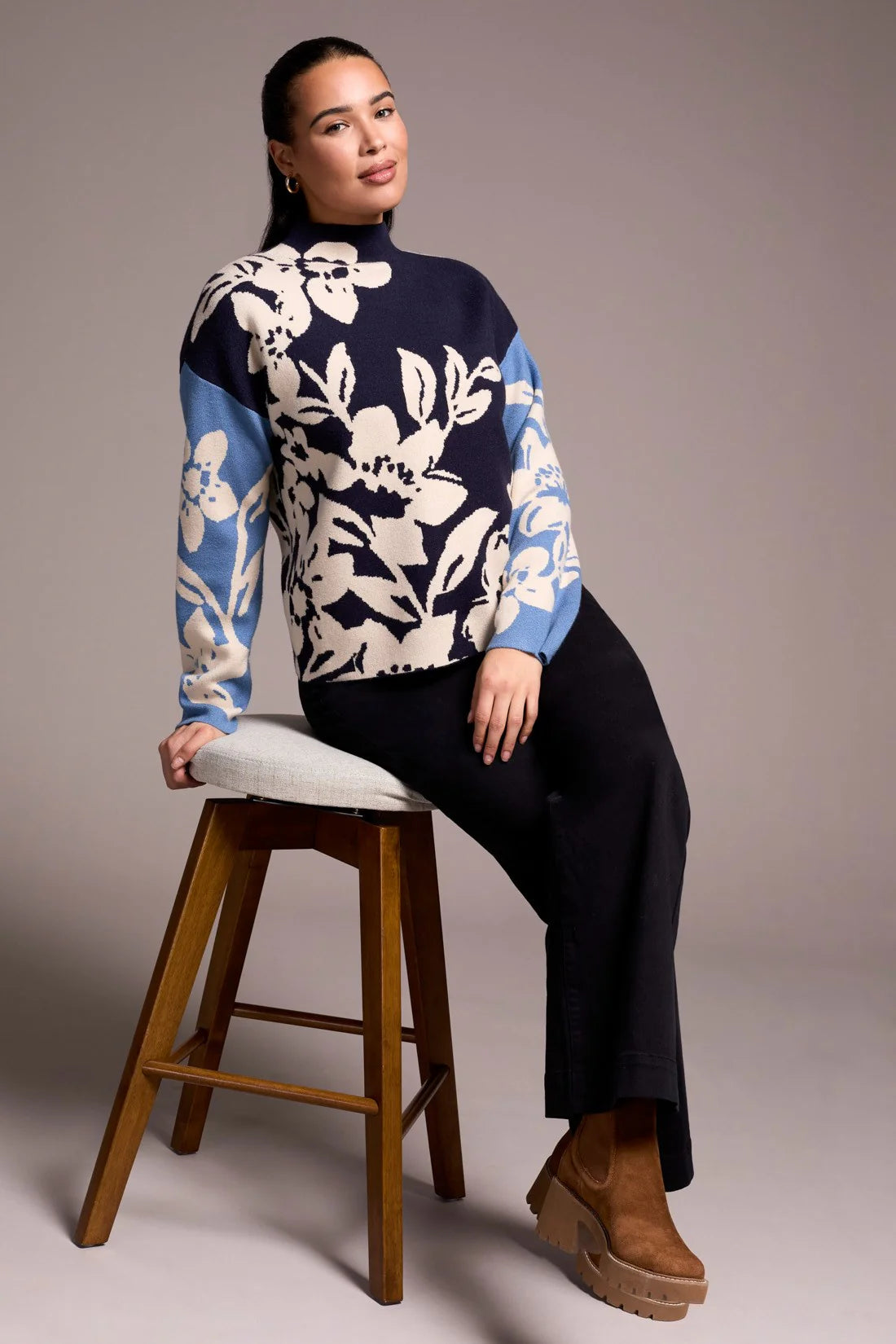 Woman wearing a floral-patterned sweater sitting on a wooden stool against a plain background
