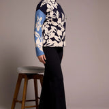 Woman wearing a blue and white floral sweater with black pants, standing next to a wooden stool against a plain background.