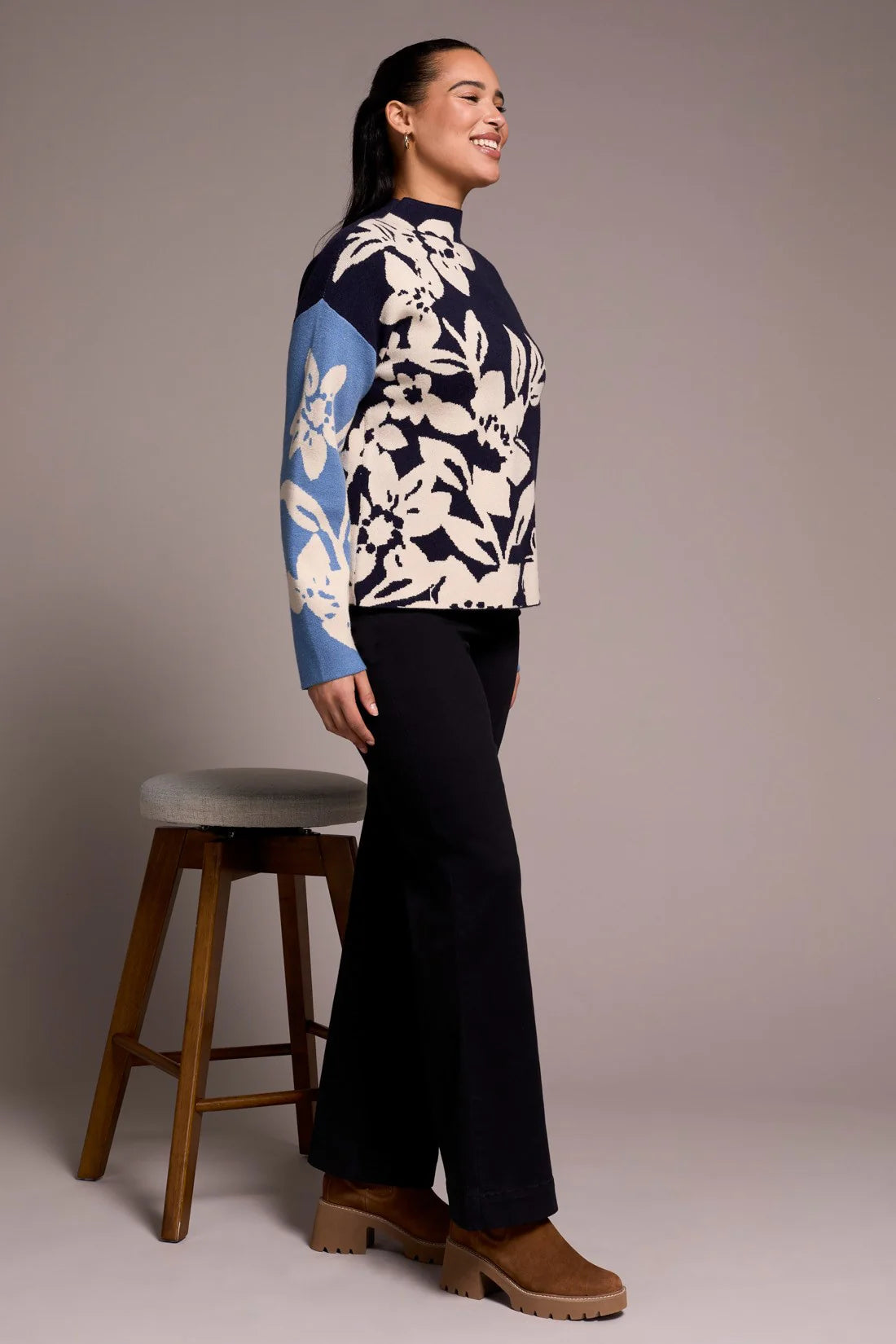 Woman wearing a blue and white floral sweater with black pants, standing next to a wooden stool against a plain background.