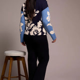 Person wearing a floral-patterned sweater with blue sleeves, standing next to a wooden stool against a plain background.