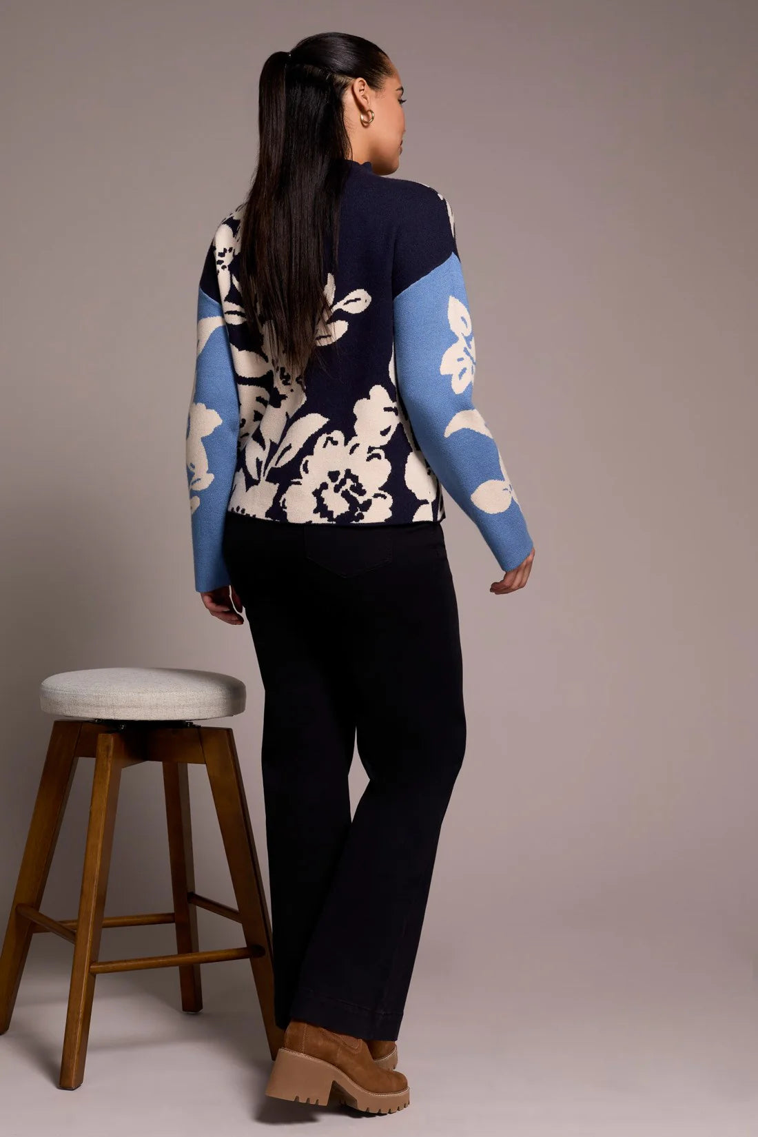 Person wearing a floral-patterned sweater with blue sleeves, standing next to a wooden stool against a plain background.