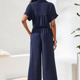 Person wearing a navy blue jumpsuit standing in a room with a white wall and plant.