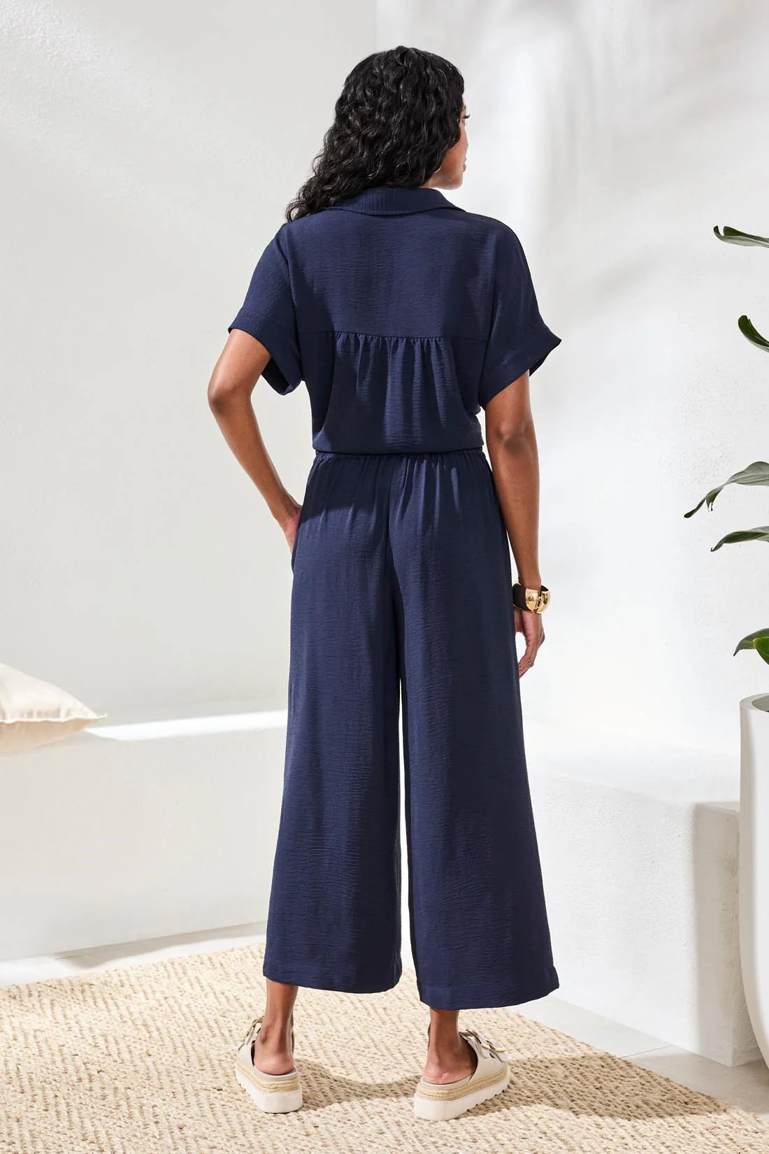 Person wearing a navy blue jumpsuit standing in a room with a white wall and plant.