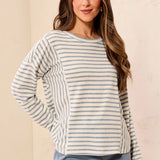 A woman wearing a long-sleeve crewneck top with horizontal blue and beige stripes, paired with light blue pants, standing against a textured beige wall.