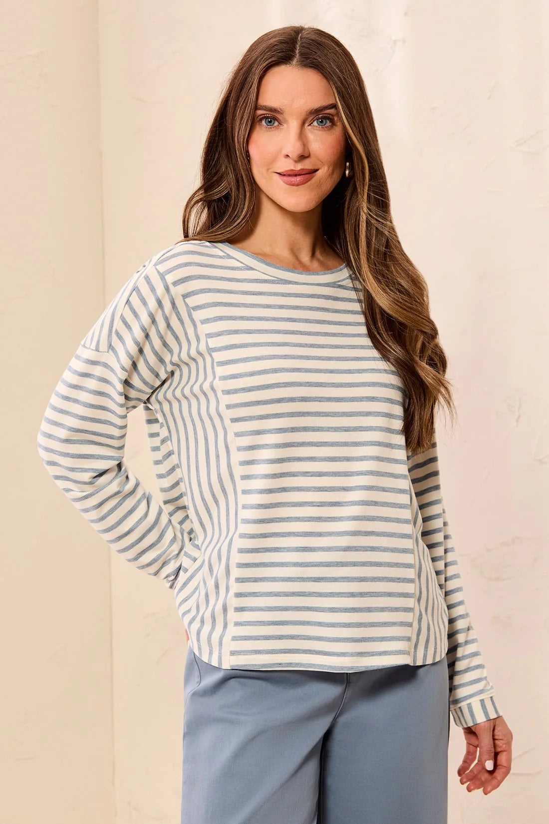 A woman wearing a long-sleeve crewneck top with horizontal blue and beige stripes, paired with light blue pants, standing against a textured beige wall.