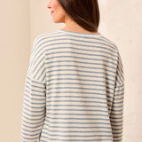 A woman shown from the back wearing a long-sleeve striped top with thin blue and off-white horizontal stripes, paired with light blue pants, set against a neutral cream background.