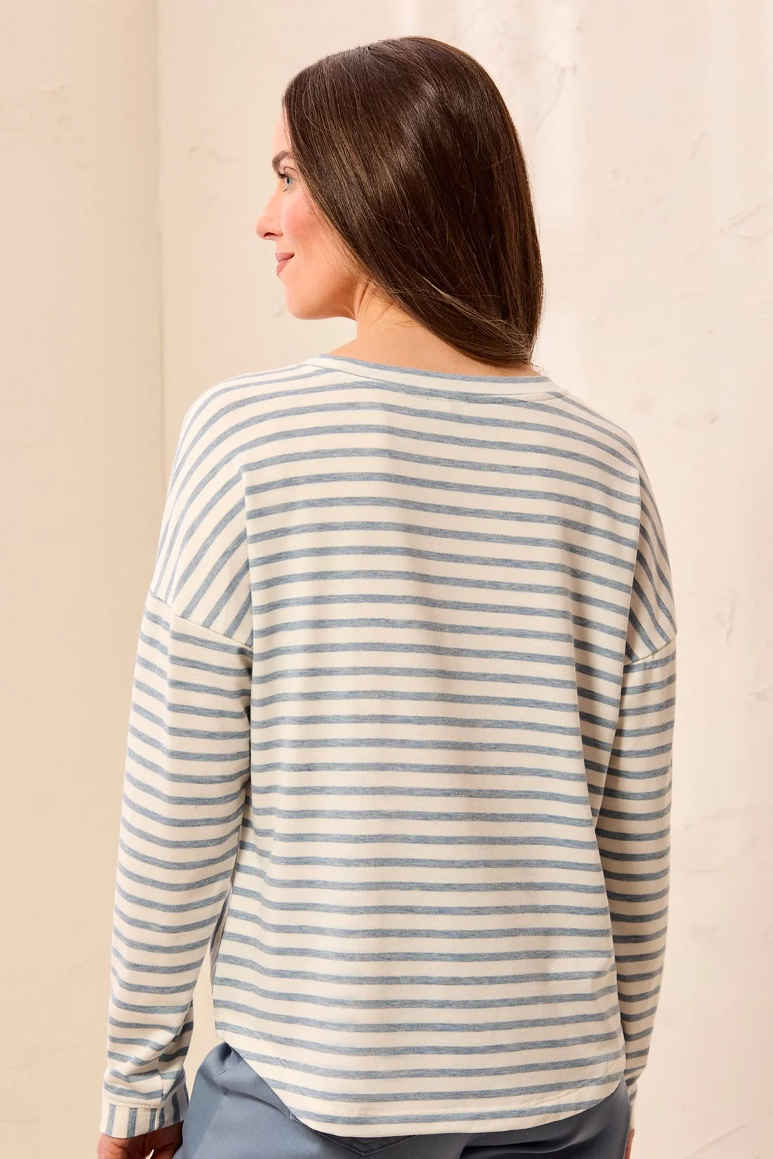 A woman shown from the back wearing a long-sleeve striped top with thin blue and off-white horizontal stripes, paired with light blue pants, set against a neutral cream background.