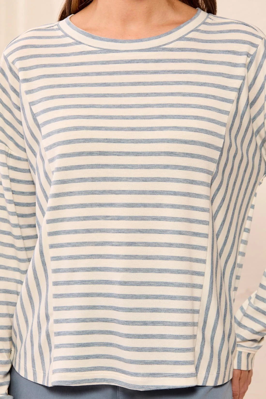 A close-up of a person wearing a long-sleeve crewneck top with thin horizontal blue stripes on a white or off-white base.