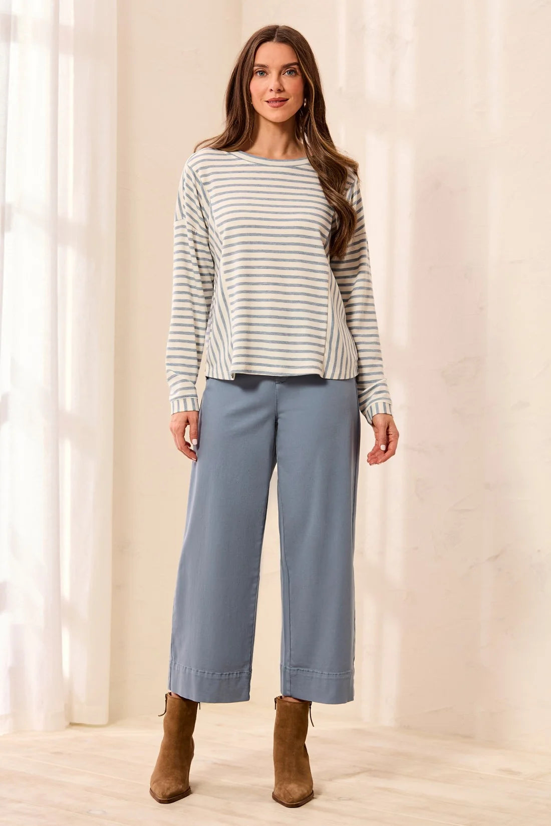 A woman modeling a long-sleeve striped knit top and wide-leg cropped pants in a softly lit indoor setting with curtains and a light floor.