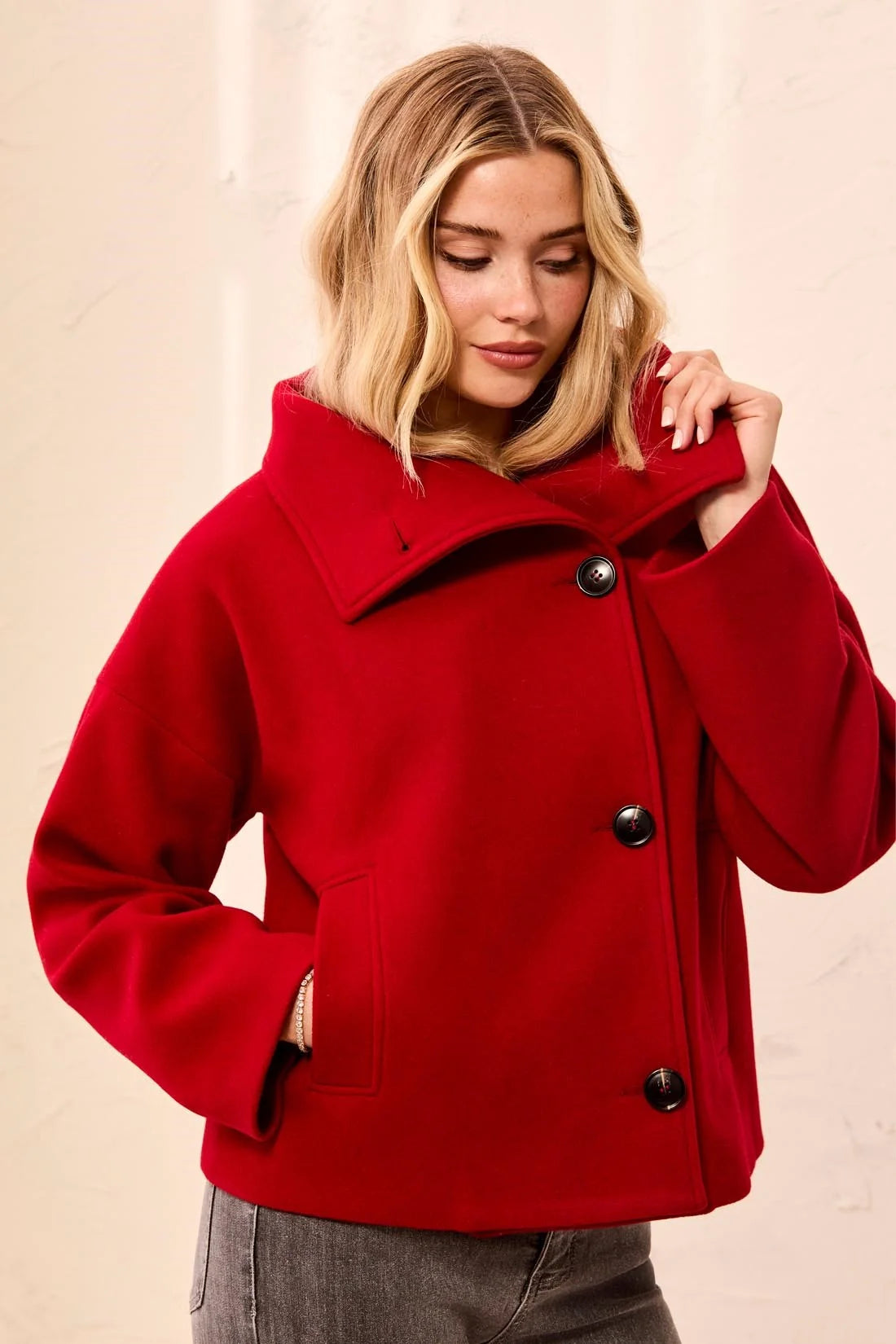 A woman wearing a bright red wool-blend coat with a wide collar and three dark buttons, paired with gray jeans, against a light beige textured wall.