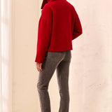 A woman shown from the back wearing a bright red cropped turtleneck sweater, gray skinny jeans, and metallic silver ankle boots, standing against a warm off-white textured wall.