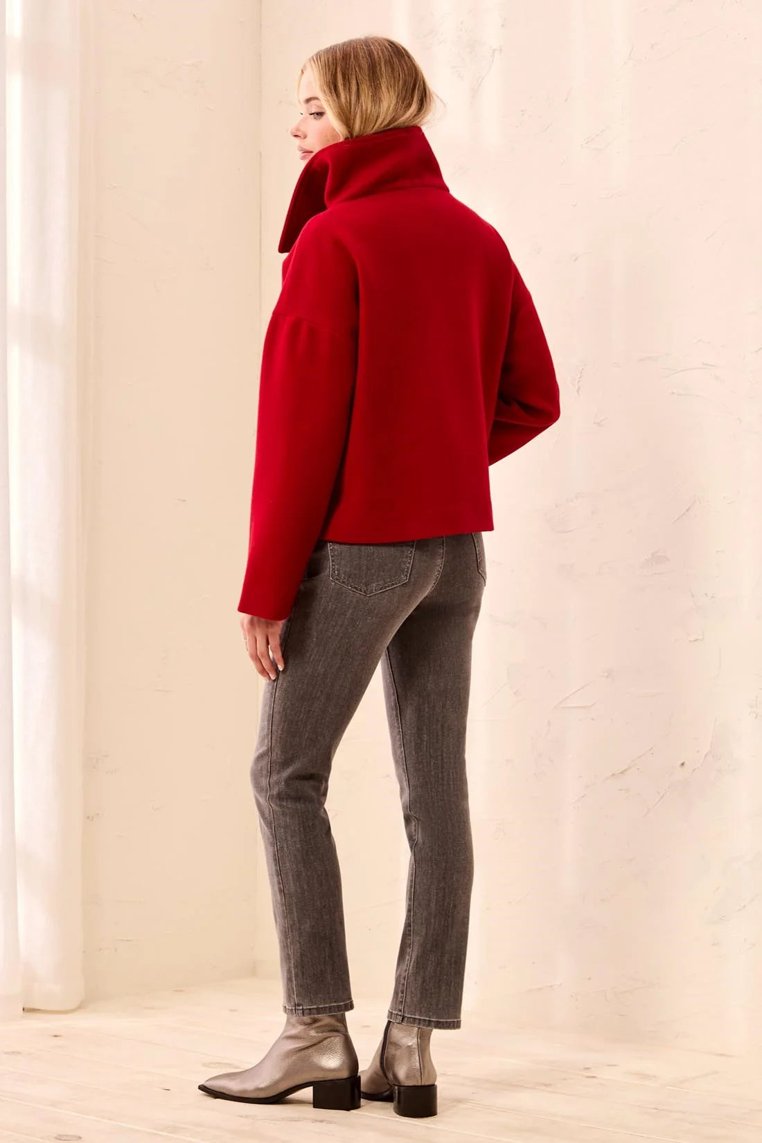A woman shown from the back wearing a bright red cropped turtleneck sweater, gray skinny jeans, and metallic silver ankle boots, standing against a warm off-white textured wall.