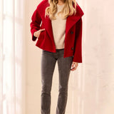A woman modeling a bright red double-breasted coat over a cream knit sweater, paired with gray skinny jeans and taupe ankle boots, standing in a brightly lit indoor setting with white curtains.