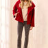 A woman modeling a bright red double-breasted coat over a cream knit sweater, paired with gray skinny jeans and taupe ankle boots, standing in a brightly lit indoor setting with white curtains.