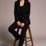 Woman in a black suit sitting on a wooden stool against a plain background