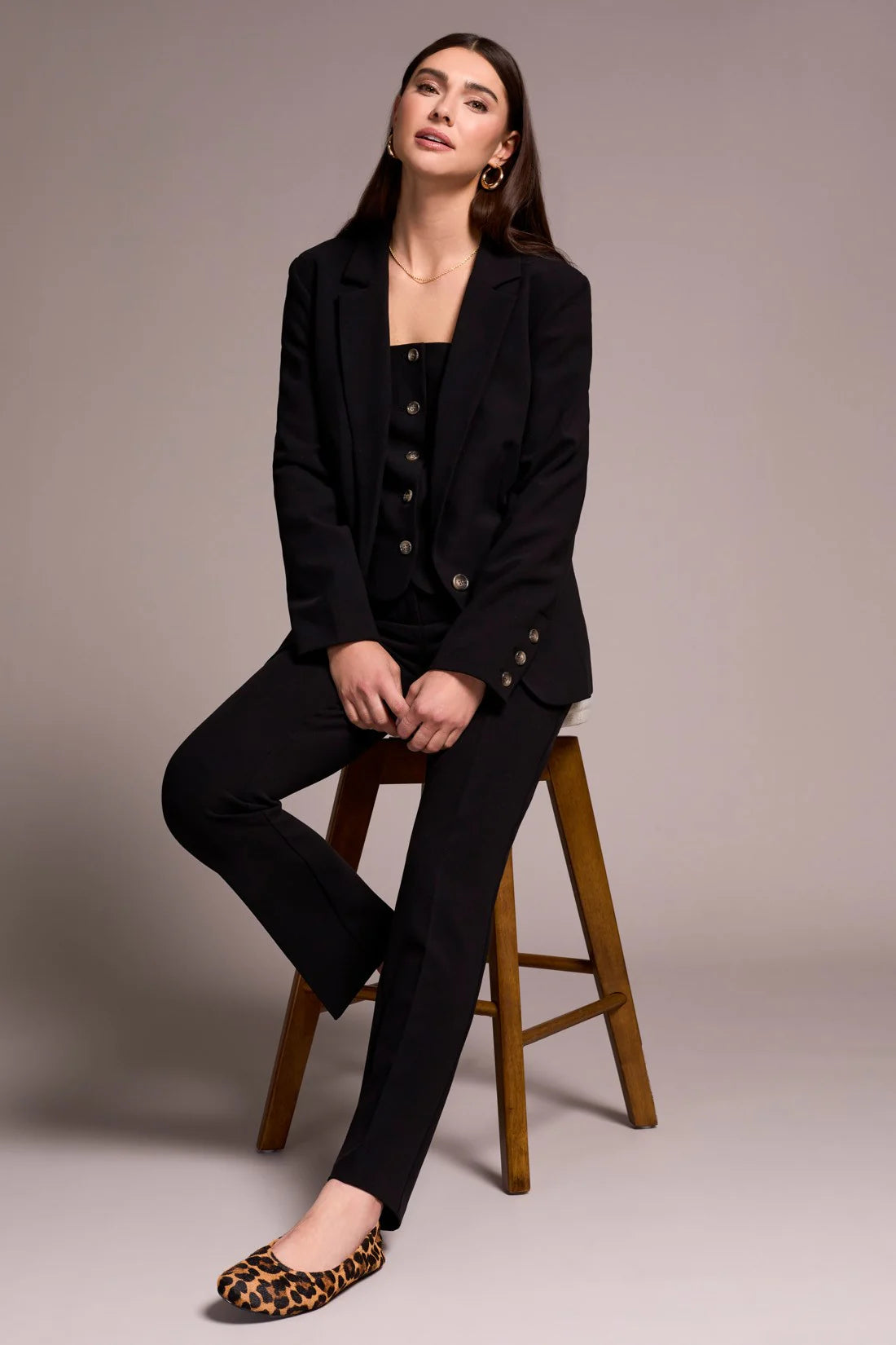 Woman in a black suit sitting on a wooden stool against a plain background