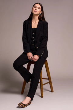 Woman in a black suit sitting on a wooden stool against a plain background
