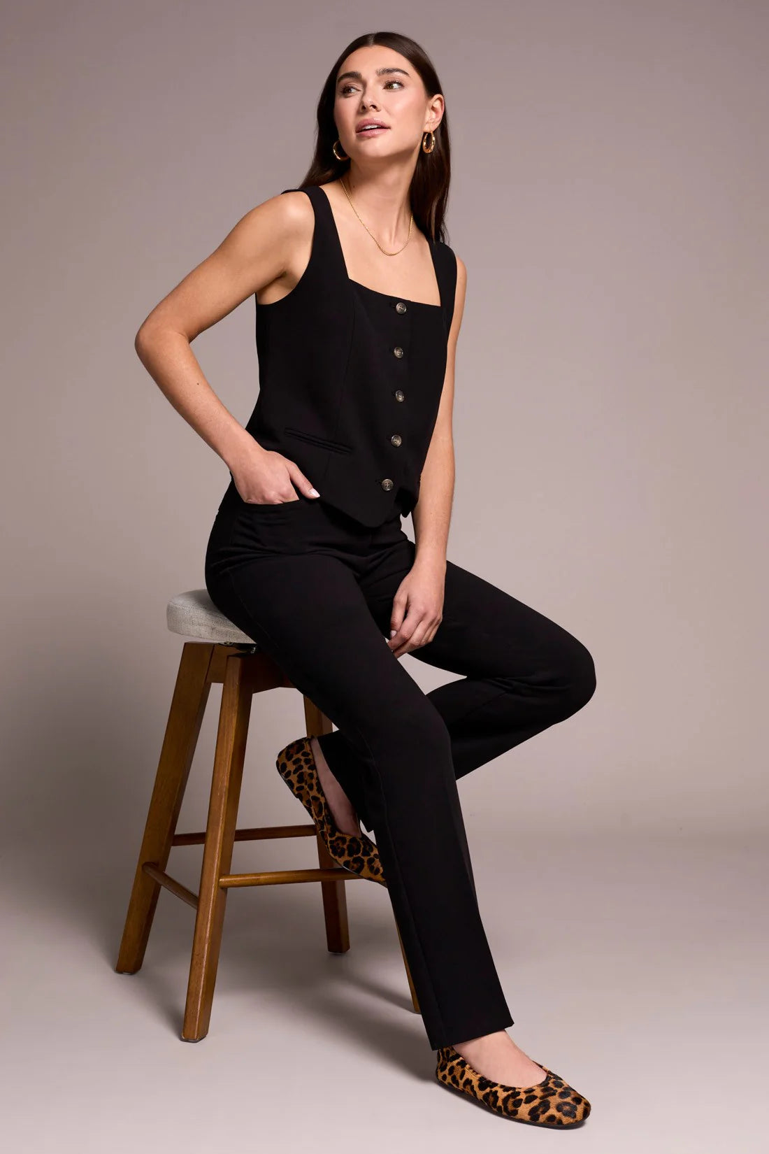 Woman sitting on a wooden stool wearing a black sleeveless top and pants with leopard print shoes.