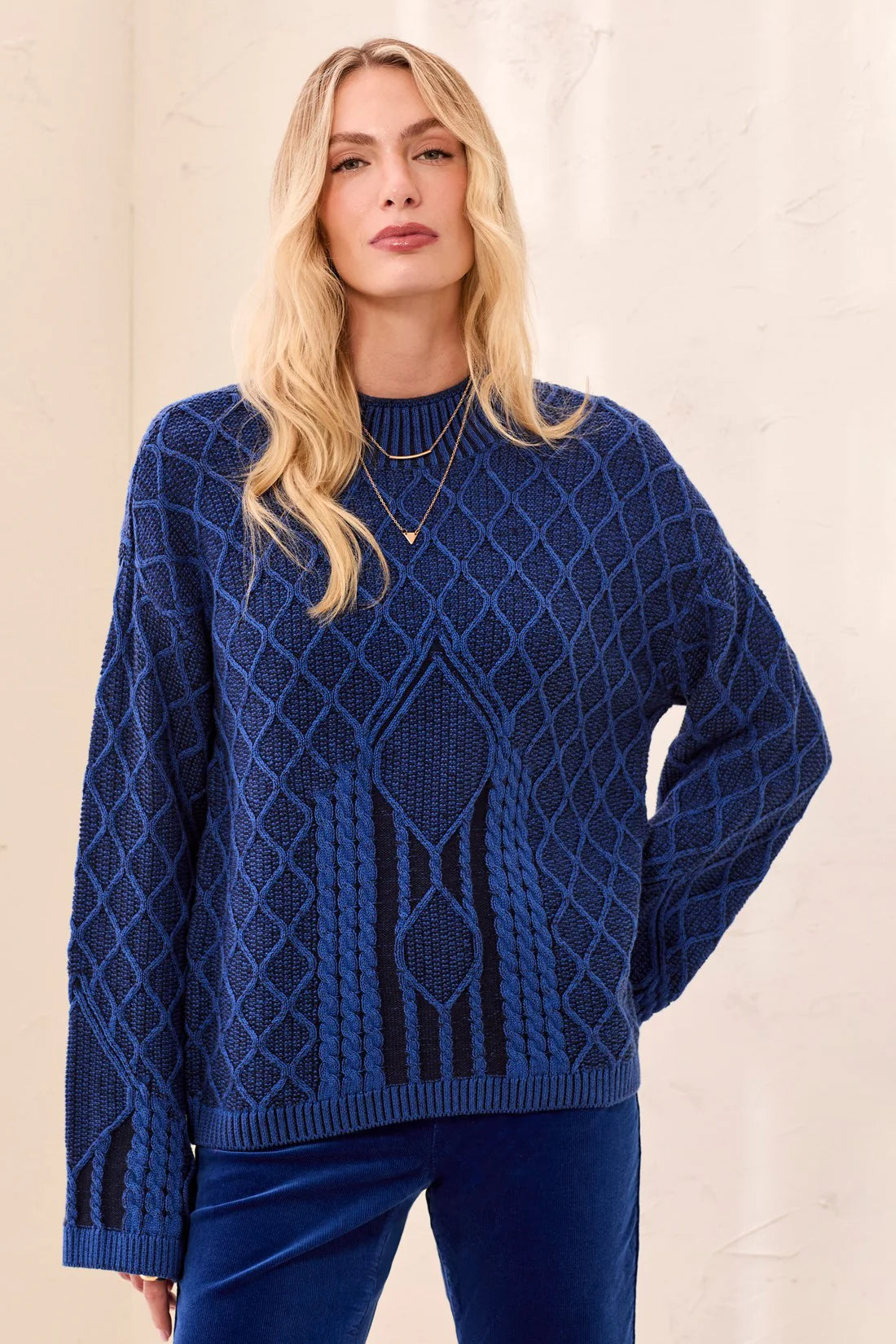 A woman wearing a textured royal blue cable-knit sweater with a diamond pattern, paired with matching blue pants, set against a neutral cream background.
