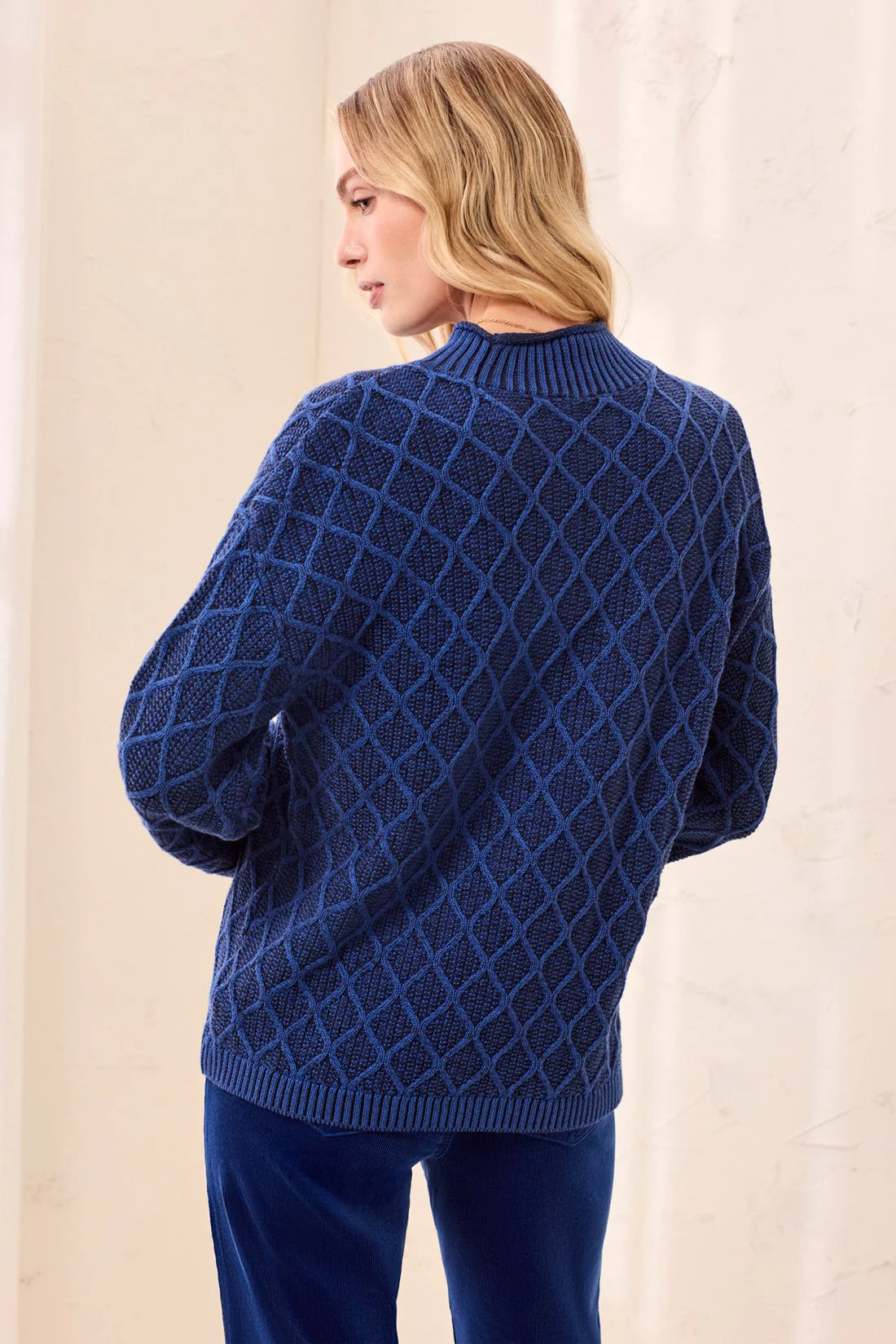 A woman shown from the back wearing a navy blue cable-knit sweater with a diamond lattice pattern and matching navy pants, set against a neutral cream-colored backdrop.