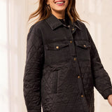 A woman wearing a black quilted jacket with a button-down front, chest flap pockets, and side pockets, photographed against a soft cream indoor background.