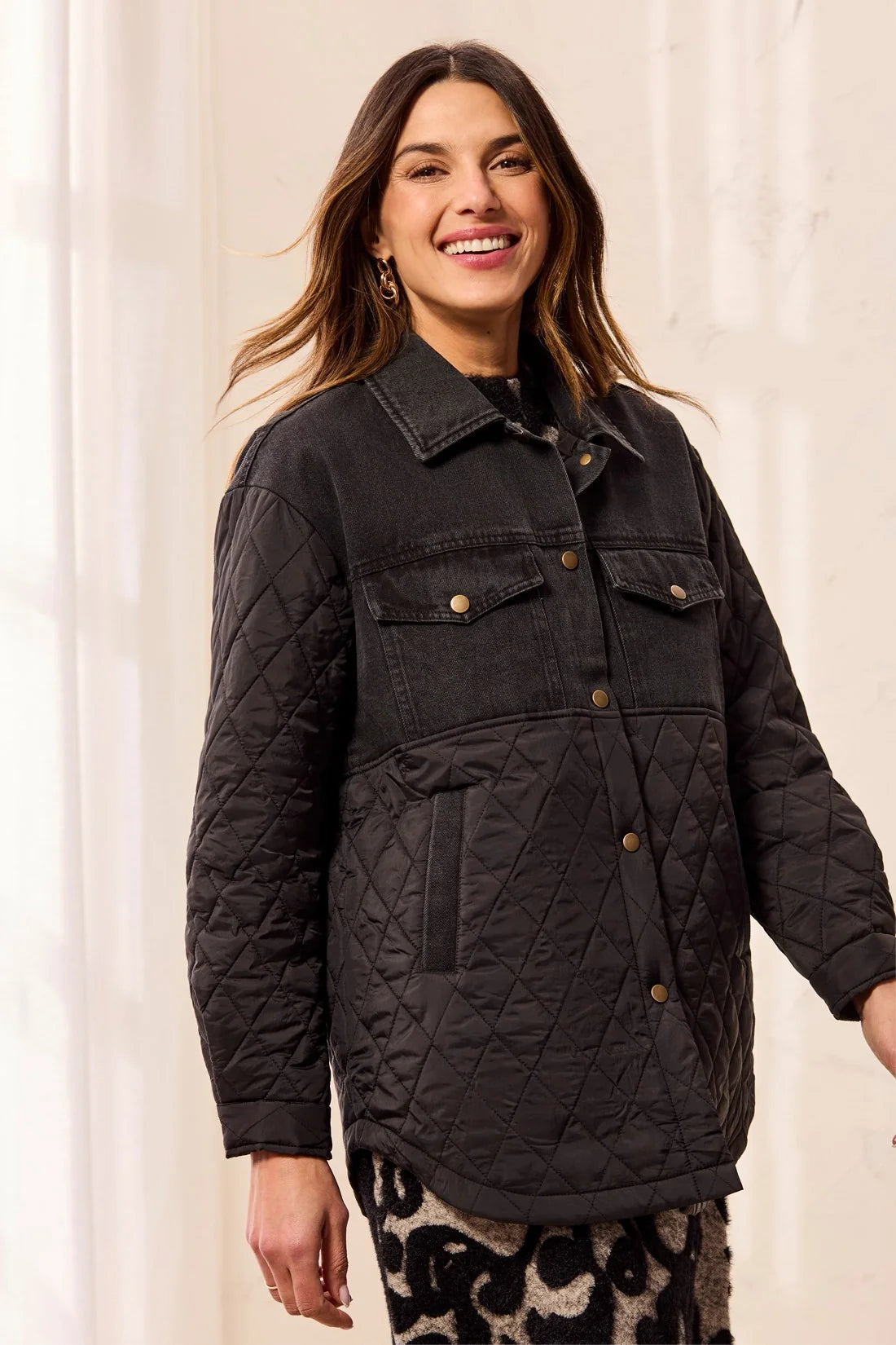 A woman wearing a black quilted jacket with a button-down front, chest flap pockets, and side pockets, photographed against a soft cream indoor background.