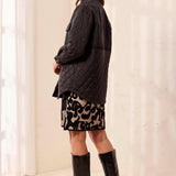 A woman wearing a black quilted jacket, a black and beige leopard-print skirt, and black knee-high boots, standing in profile against a light cream curtain backdrop.