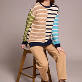 Woman sitting on a stool wearing a colorful striped sweater and beige pants.