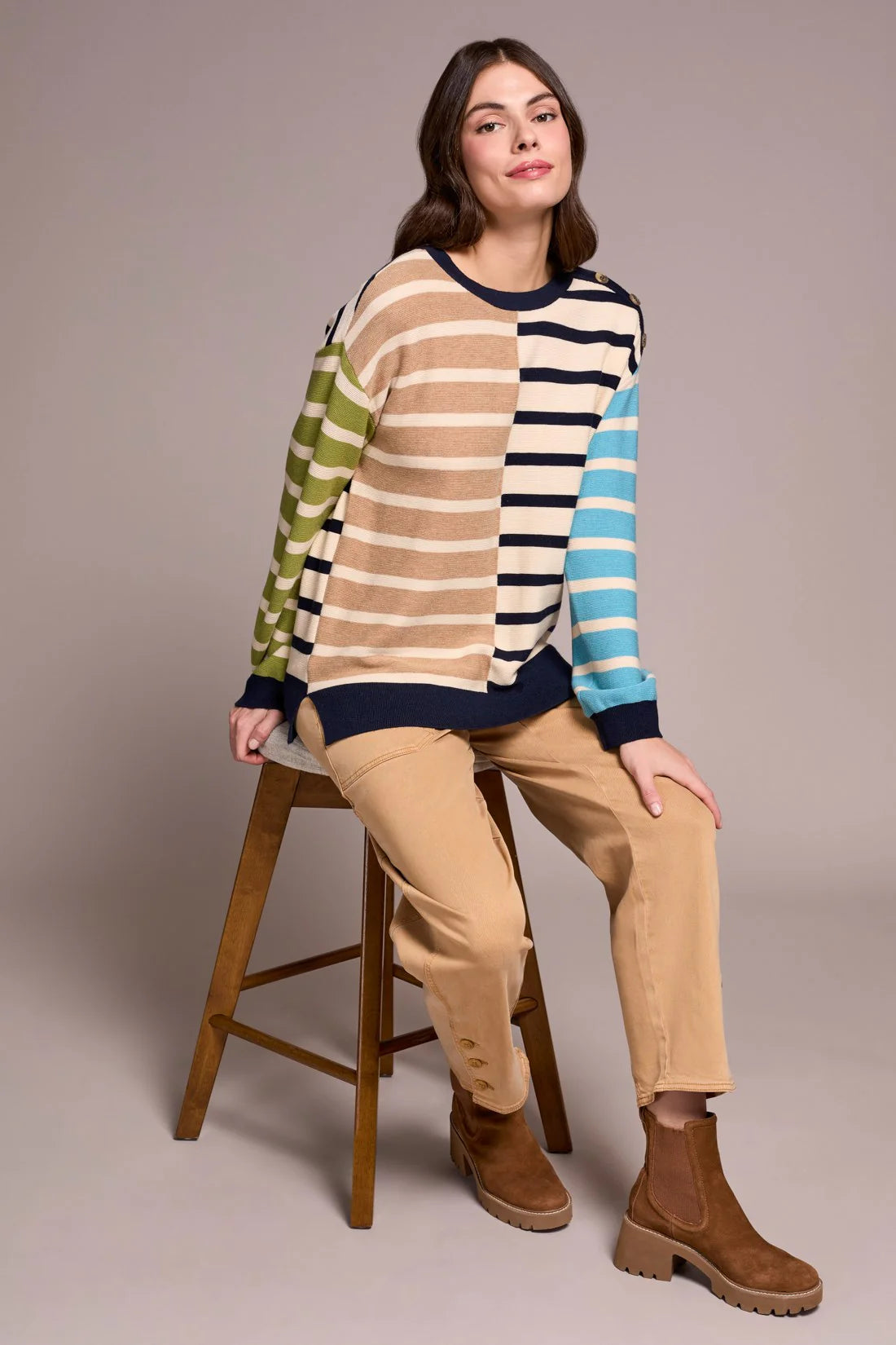 Woman sitting on a stool wearing a colorful striped sweater and beige pants.