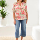 Woman wearing a floral blouse and blue jeans standing in a bright room with a plant.