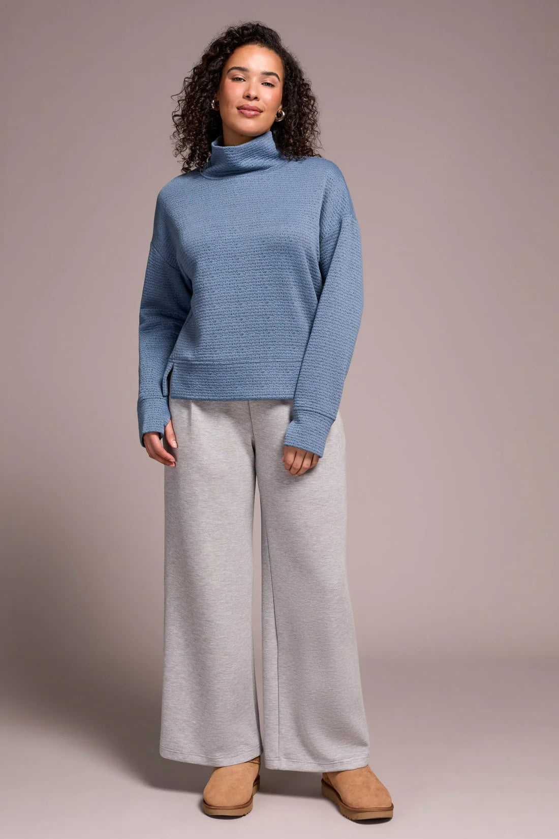 Woman wearing a blue sweater and light gray pants against a plain background