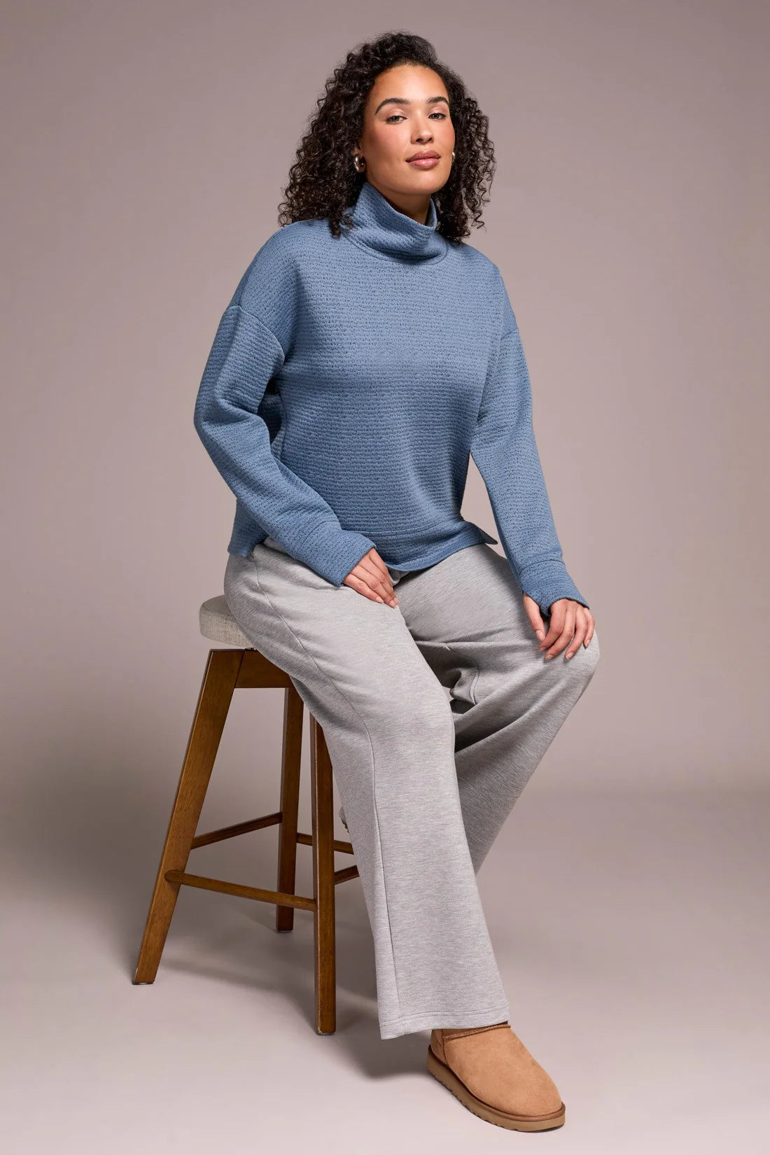 Woman wearing a blue turtleneck sweater and gray pants sitting on a wooden stool against a plain background