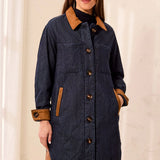 A woman wearing a dark navy blue quilted coat with contrasting brown collar, cuffs, and pocket trims, paired with brown pants, set against a soft beige curtain background.