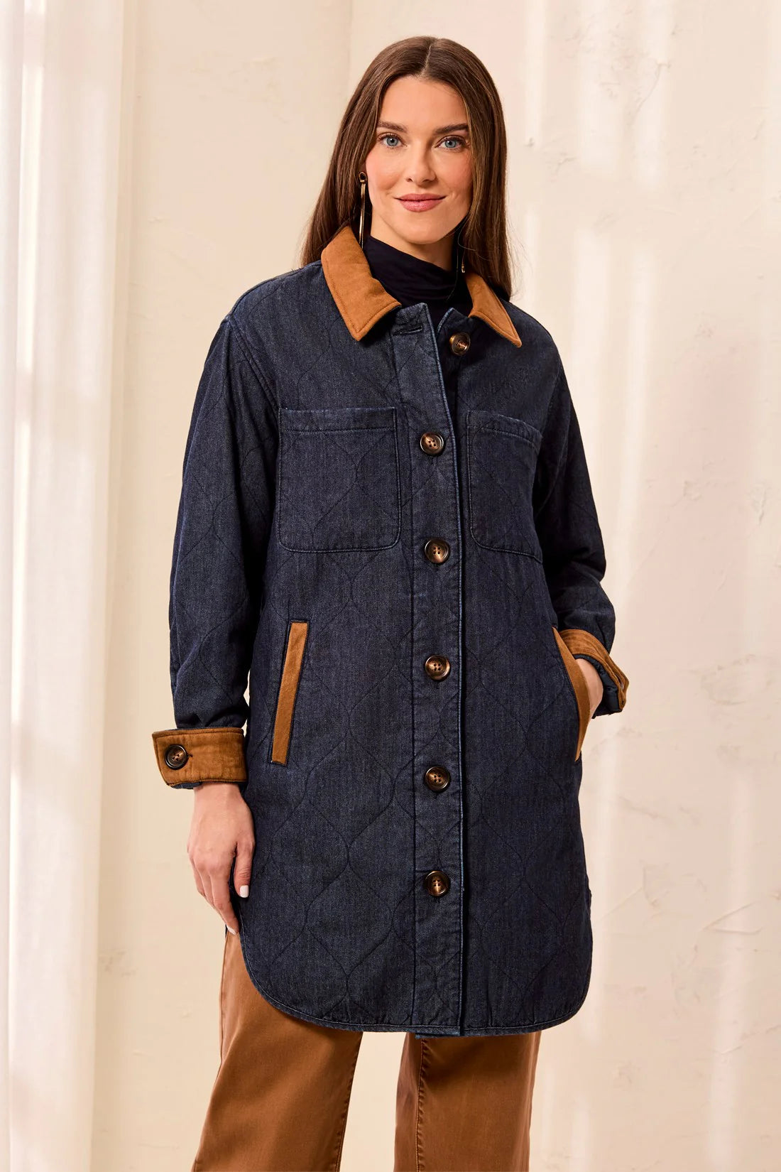A woman wearing a dark navy blue quilted coat with contrasting brown collar, cuffs, and pocket trims, paired with brown pants, set against a soft beige curtain background.