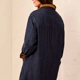 A woman shown from the back wearing an oversized dark indigo denim shirt jacket with a brown suede collar, set against a warm beige textured wall.