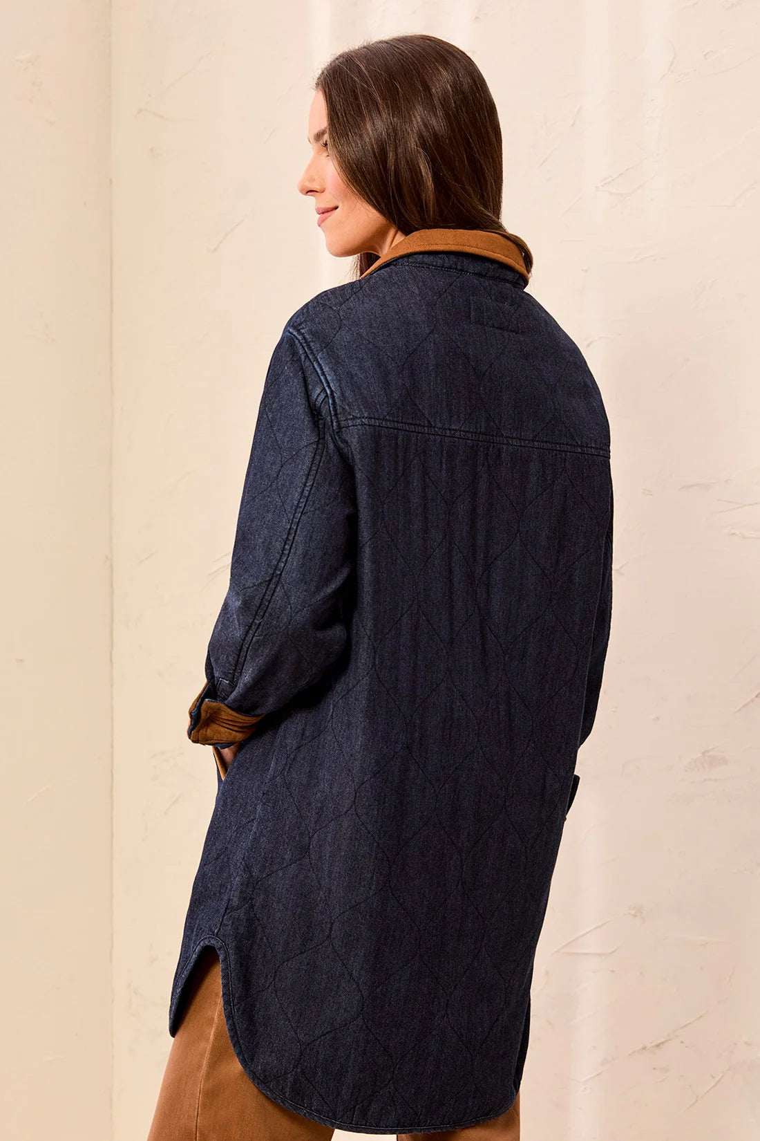 A woman shown from the back wearing an oversized dark indigo denim shirt jacket with a brown suede collar, set against a warm beige textured wall.