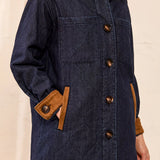 A close-up of a person wearing a dark blue quilted coat with contrasting tan collar, cuffs, and pocket trim, featuring large brown buttons.