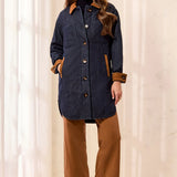 A woman modeling a dark navy quilted coat with brown suede-like trim, paired with brown wide-leg pants and matching brown ankle boots, standing in a bright indoor setting with white curtains.
