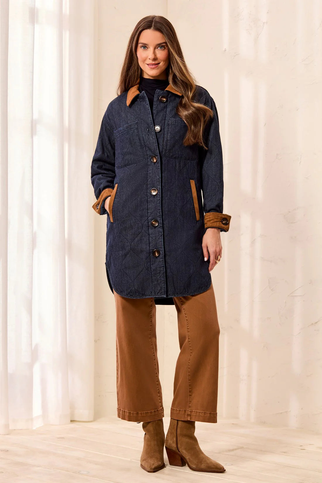 A woman modeling a dark navy quilted coat with brown suede-like trim, paired with brown wide-leg pants and matching brown ankle boots, standing in a bright indoor setting with white curtains.