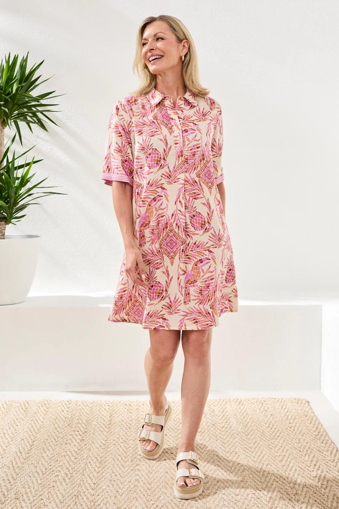 Woman wearing a pink floral dress standing in a bright room with a plant in the background.