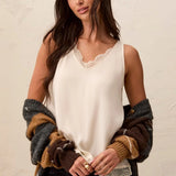 A woman wearing a cream sleeveless lace-trimmed camisole top, styled with a brown and gray knit cardigan draped off her shoulders and dark jeans, posing against a textured off-white wall.