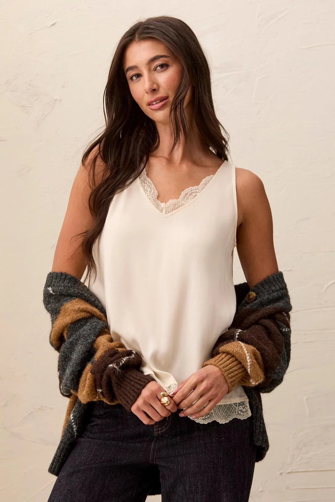 A woman wearing a cream sleeveless lace-trimmed camisole top, styled with a brown and gray knit cardigan draped off her shoulders and dark jeans, posing against a textured off-white wall.