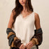 A woman wearing a cream sleeveless lace-trimmed camisole top, styled with a brown and gray knit cardigan draped off her shoulders and dark jeans, posing against a textured off-white wall.