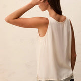 A woman shown from the back wearing a sleeveless cream-colored top with a delicate lace hem, paired with dark jeans, against a textured off-white wall.