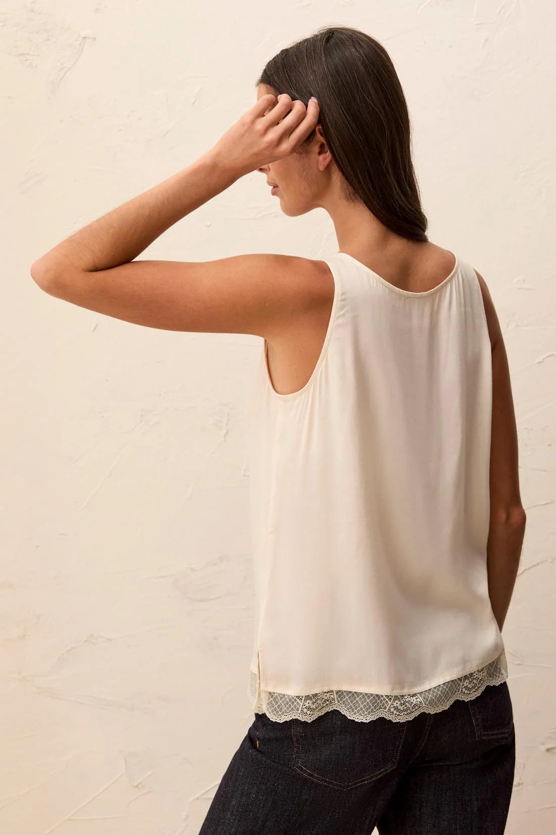 A woman shown from the back wearing a sleeveless cream-colored top with a delicate lace hem, paired with dark jeans, against a textured off-white wall.