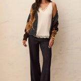 A woman modeling a bohemian-style outfit consisting of a cream lace-trimmed camisole, dark-wash wide-leg jeans with rolled cuffs, a brown and green patterned cardigan, and black ankle boots, standing against a textured beige wall.