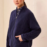 A woman wearing a navy blue quilted zip-up jacket over a patterned top and matching patterned skirt, standing against a light beige textured wall.