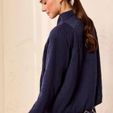 A woman wearing a dark navy blue textured long-sleeve jacket and a patterned skirt, shown from a side-back angle against a light beige wall.